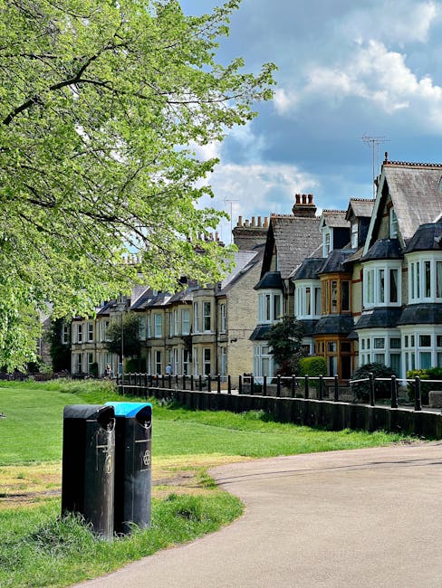 A row of traditional terraced houses with bay windows, slate-tiled roofs, and brick chimneys, situated behind a small, neatly maintained grassy area. In the foreground, two black cylindrical rubbish b